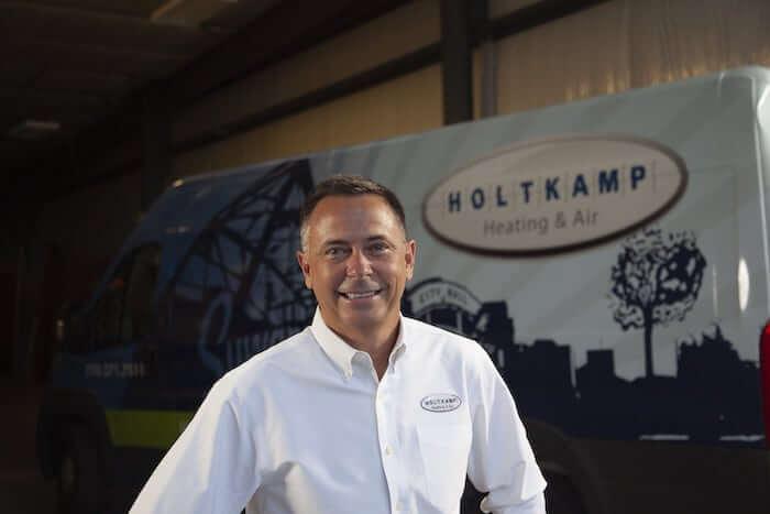 Matthew Holtkamp from Holtkamp Heating & Air stands in front of a sign displaying the company logo.