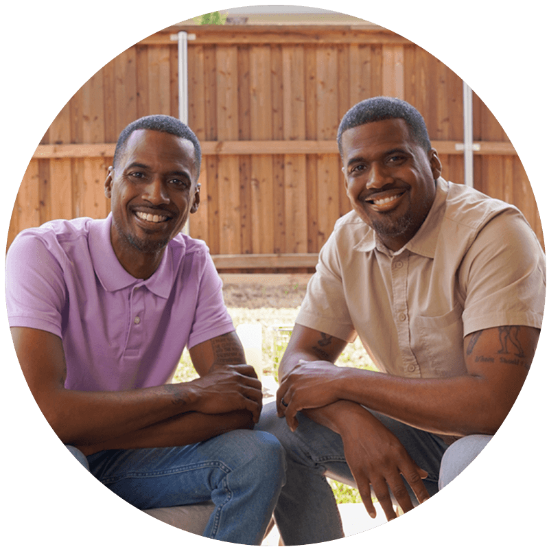 Two African-American men, one in a pink shirt and the other in a tan shirt, are seated at a patio table.